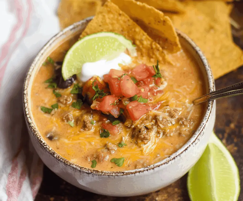 Creamy cheesy ground beef taco soup in a bowl topped with shredded cheese, fresh herbs, and crispy tortilla chips